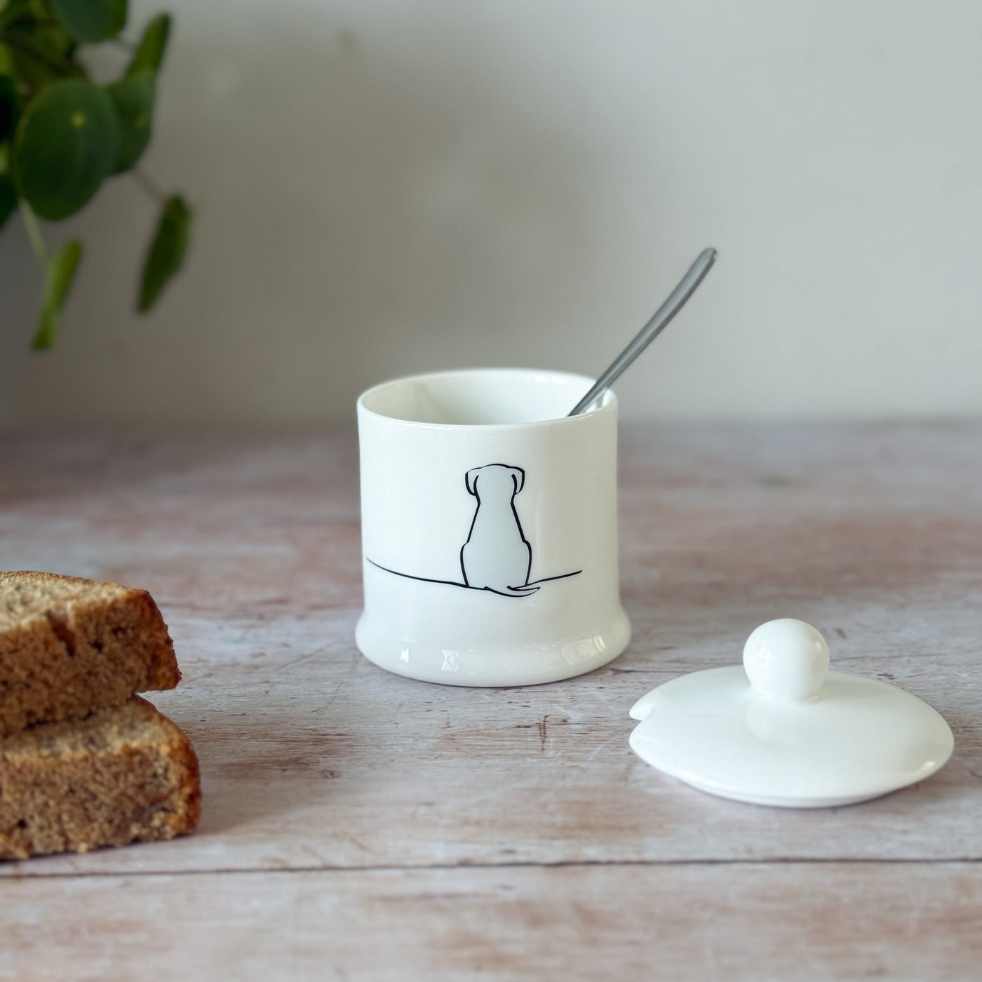 White fine bone china jam pot with Sitting Dog illustration on a wooden surface with cake and a plant.