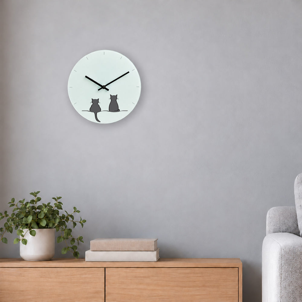 A minimalist round glass wall clock featuring Two Cats Together by Jin Designs mounted on a wall above a wooden sideboard with a potted plant and books.