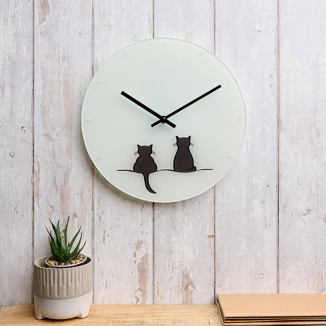 A round glass wall clock featuring Two Cats Together Limited Edition by Jin Designs. The clock is mounted on a whitewashed wood plank wall above a wooden surface with a small potted succulent and a stack of cards nearby.