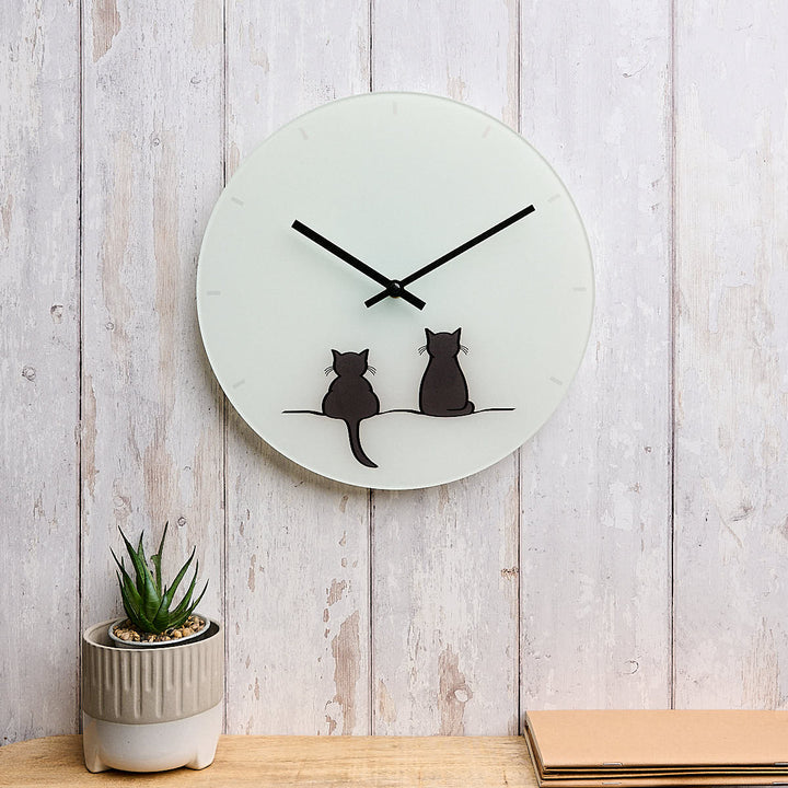 A round glass wall clock featuring Two Cats Together Limited Edition by Jin Designs. The clock is mounted on a whitewashed wood plank wall above a wooden surface with a small potted succulent and a stack of cards nearby.