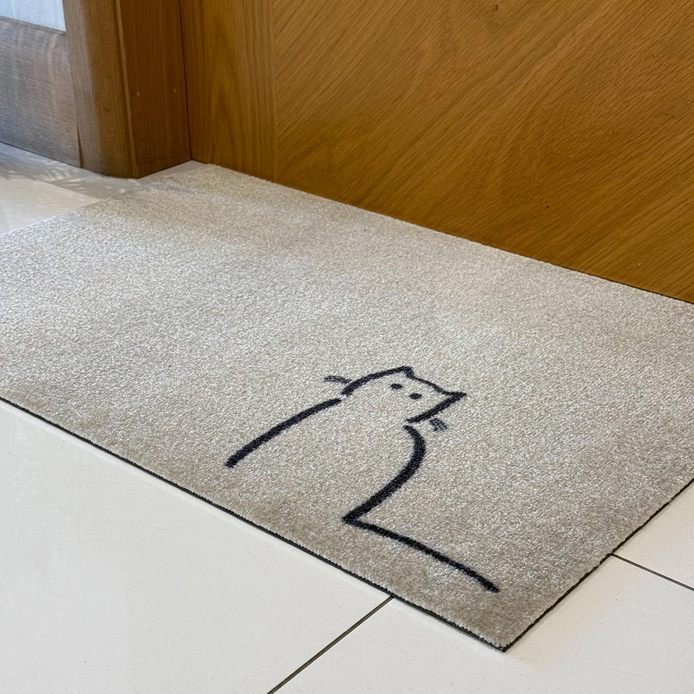 Waiting Cat Doormat by Jin Designs - a rectangular beige doormat with a simple black line drawing of a cat, placed on a white tiled floor in front of a wooden door.