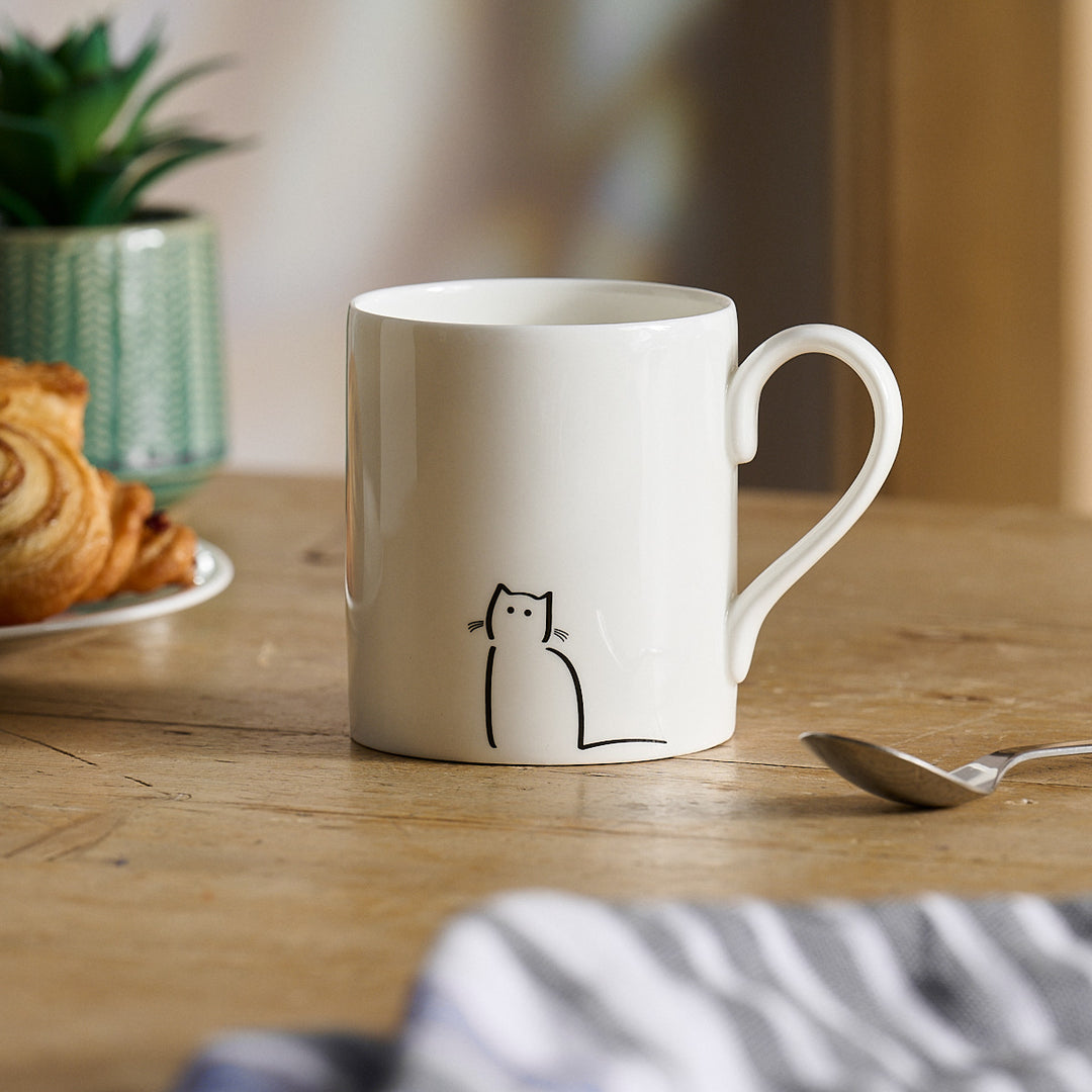 Waiting Cat White mug with a cat illustration on a wooden table with a croissant and plant in the background