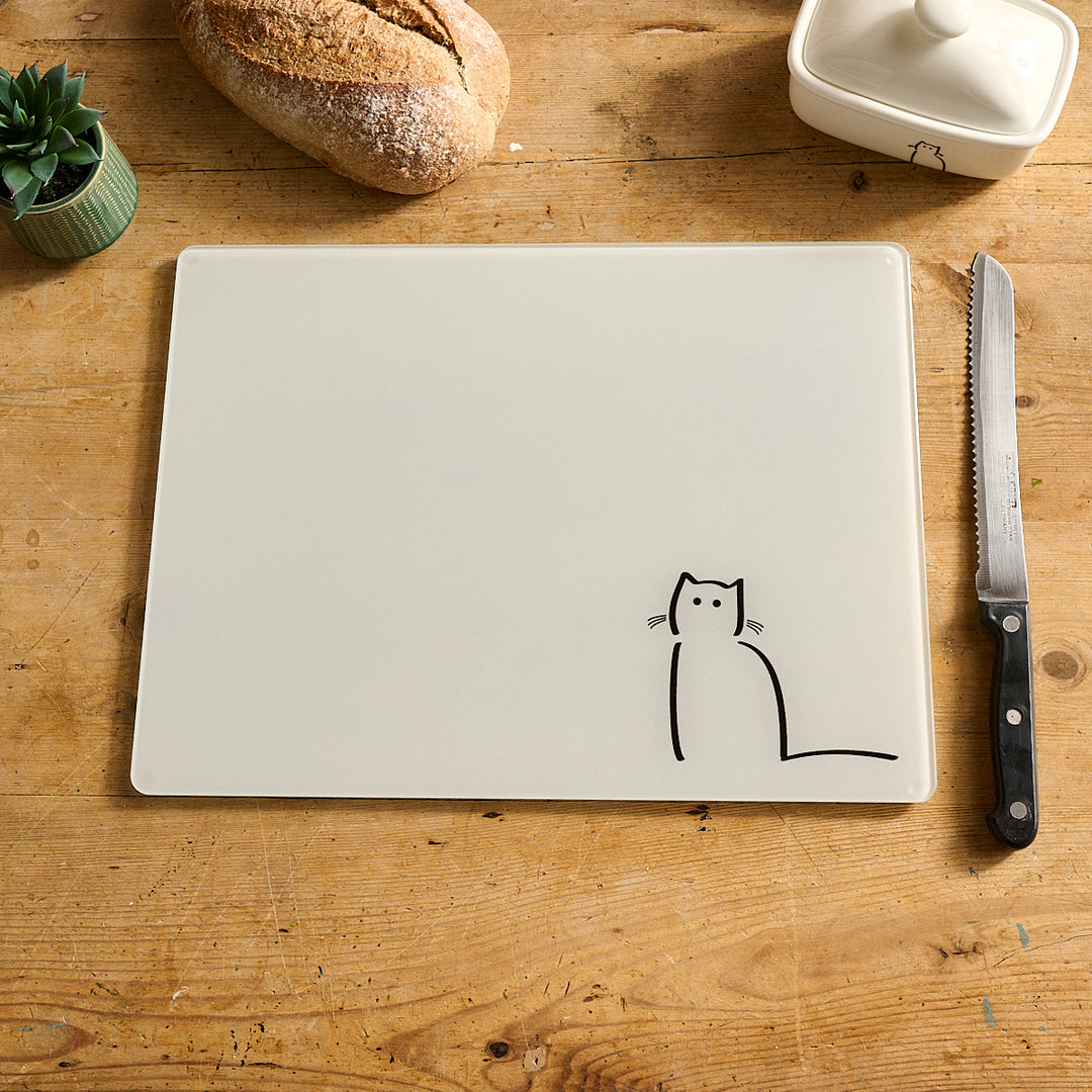 Waiting Cat Glass Worktop Saver with a black cat drawing on a wooden table with bread and a knife.