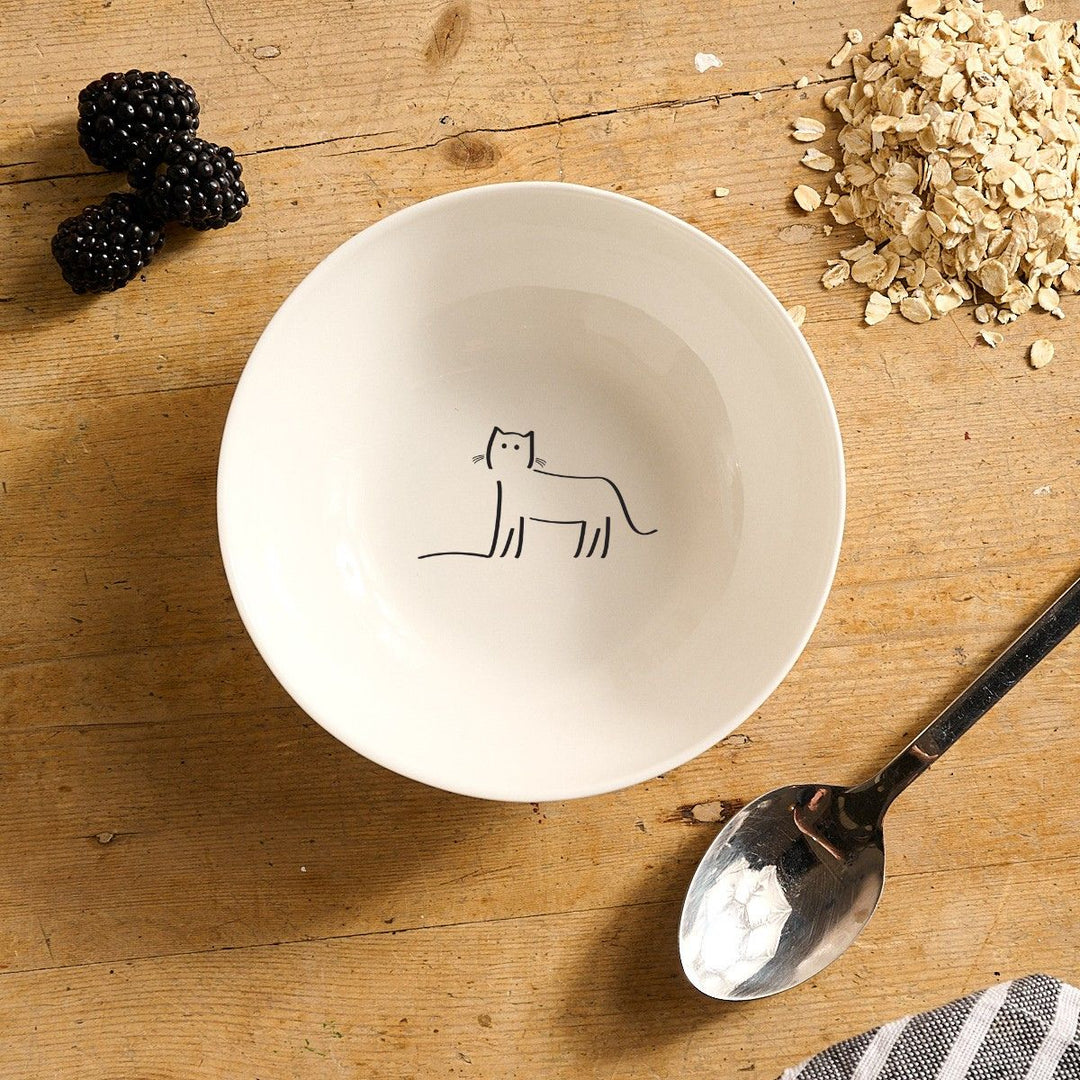 White bowl with Watching Cat design on a wooden surface with blackberries, oats, and a spoon.