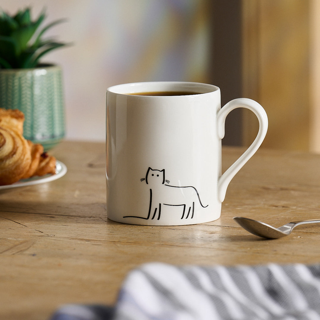 Watching Cat Mug with a cat illustration on a wooden table with coffee, croissant, and spoon.