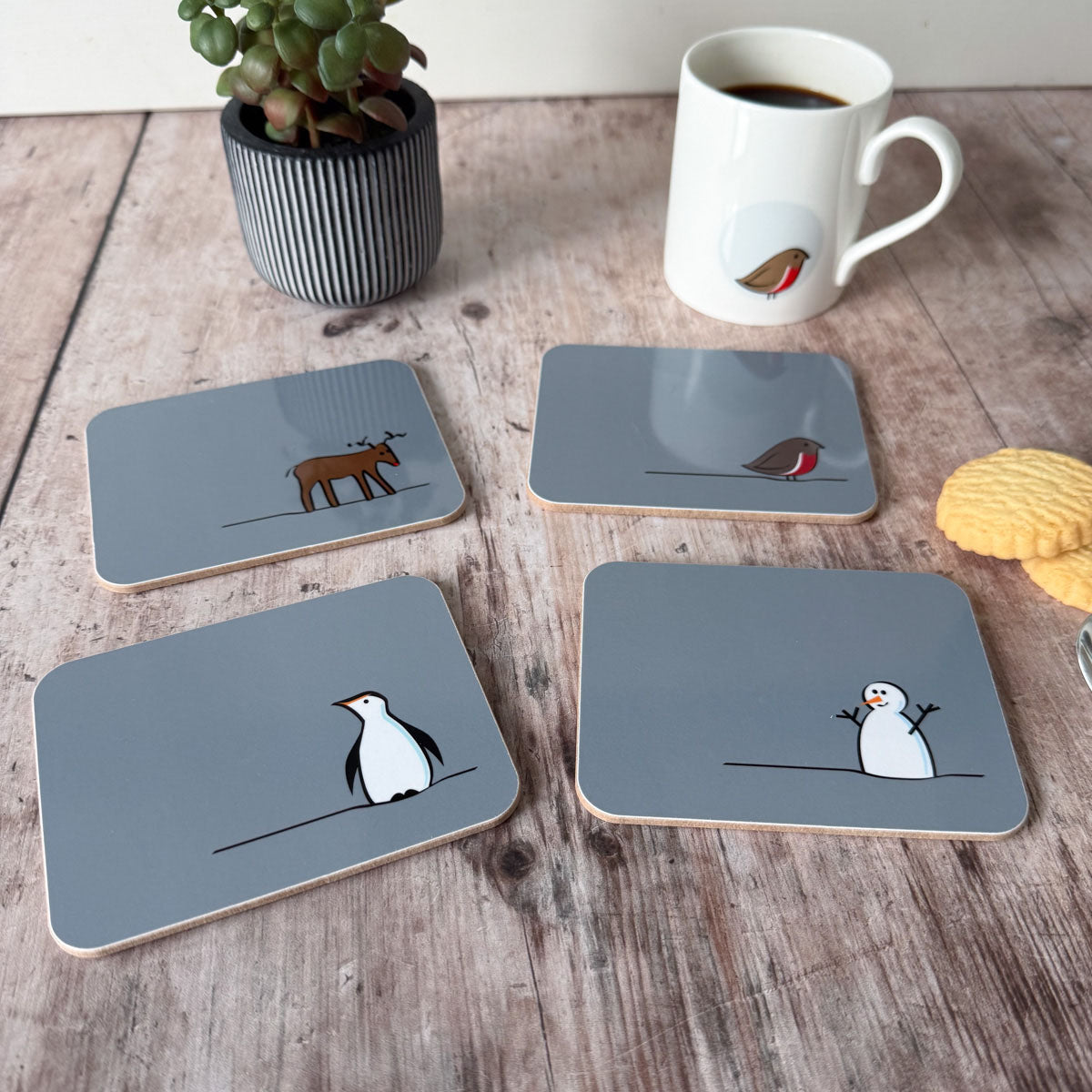 Set of four Winter Collection coasters with animal illustrations on a wooden surface with a mug and plant.