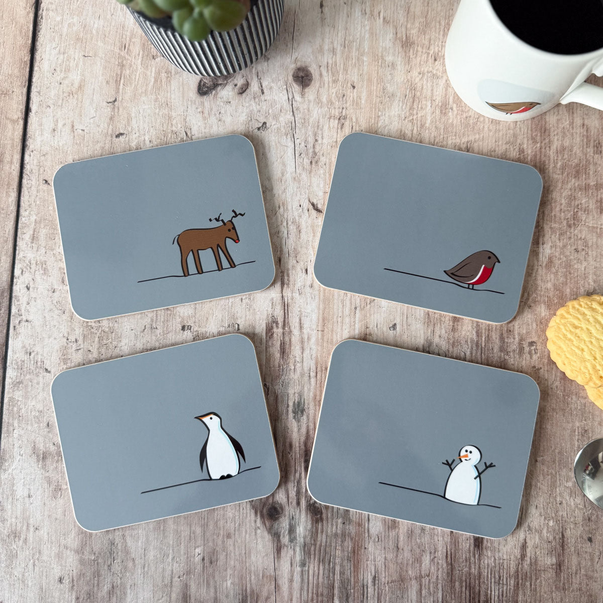 Four Winter Collection coasters with animal illustrations on a wooden surface with a mug and plant.
