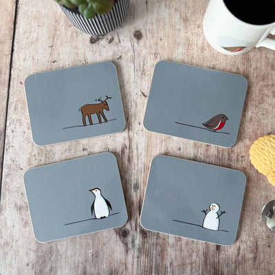 Four Winter Collection coasters with animal illustrations on a wooden surface with a mug and plant.